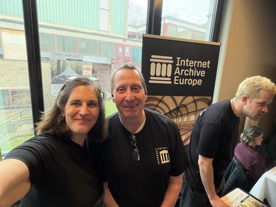 A man and a woman pose for a selfie as another man interacts with the public at the Internet Archive Europe stand at FOSDEM in Brussels