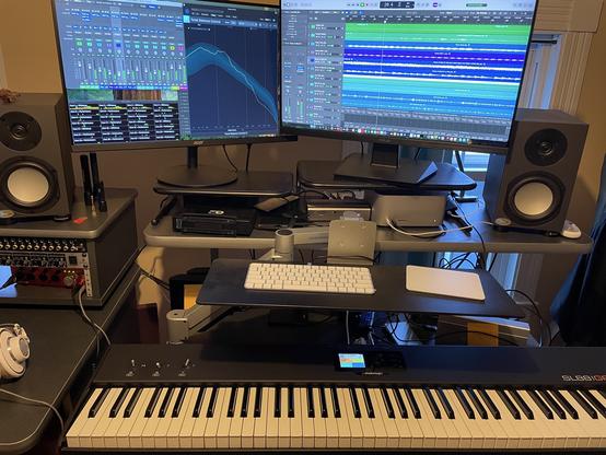 A dual-monitor computer screen with studio speakers on either side of them. In front of this is a 2-tiered stand with computer keyboard and trackpad on the smaller, upper one and a musical keyboard (88 keys) on the lower one.