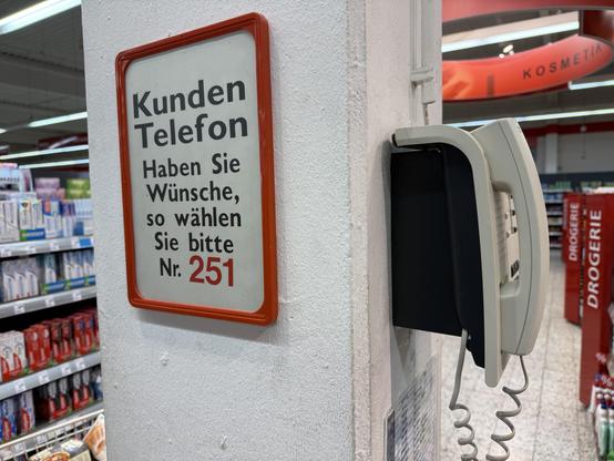 Säule in einem großen Supermarkt, an ihr hängt ein altes Telefon mit Spiralkabel. Daneben ein Schild: „ Kunden Telefon. Haben Sie Wünsche, so wählen Sie bitte Nummer 251“