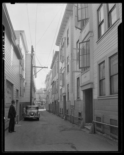 The image shows a black and white street scene from an alleyway in San Francisco, California. The architecture is characterized by narrow buildings with wooden facades and metal railings on the sides of some windows. A vintage car parked at the end of the alley catches attention due to its classic design featuring round headlights and chrome accents.

On the left side of the image, a figure appears to be walking down the street, dressed in dark clothing that blends with shadows cast by overhead wires or power lines extending from buildings above. The overall atmosphere is quiet and somewhat desolate, suggesting it could have been taken during an early morning or late afternoon when foot traffic was minimal.

The vintage car's presence suggests this photo might date back several decades ago. Overall, the image captures a moment in time within San Francisco’s Italian district known as North Beach (or "North Bean," possibly indicating historical documentation). The setting reflects urban decay and contrasts with modern-day cityscapes typically associated with bustling streets filled with people.

Additional context for understanding this photo can be found at Card Alley.