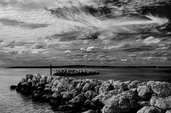 Here is a black and white breakwater photo I took in the Bahamas, off Great Abaco. These man-made structures are designed to mimic the appearance of shoals, but their job is to protect the coast from high-energy waves and storms. The pitted rocks stretch diagonally across the sea, from right to left, like a bridge over calm waves. The sky is strewn with a delicate mixture of clouds; my favorite being the wispy cirrus in the upper right that looks like a fishbone or a feathery arrow. Between the rugged rocks and soft sky, the ocean appears wrinkly, almost silken, leading to a distant line of shore. The tinest sailboat drifts on the right, facing left.

Standing before this clutch of serenity, I couldn’t help but wonder: what stories does each rock hold? Every dip, crack, and dent becomes a word. From all angles, different pages emerge. Yet, all we can read is on the surface. What dwells deeper within or beneath the tides? An ocean of libraries. These thoughts inspired another haiku in my travel journal.

clouds shape narrative
shoal rock tales
form a library