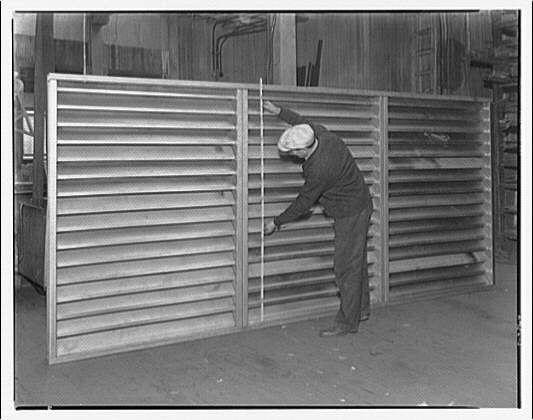 The image is a black and white photograph showing an individual working with large metal slats, which appear to be part of a construction or manufacturing process. The person is wearing work clothes including trousers, shoes, and what seems like protective gloves on both hands as they handle the metal components.

They are standing next to two vertical stacks of these slatted sheets that are likely being constructed into an enclosure or barrier system. Each stack consists of numerous horizontal lines indicating different panels within a larger unit. The background is somewhat blurred but shows signs of industrial surroundings, including various structures and possibly other equipment out of focus in the distance.

The image has historical significance as it depicts construction activities from approximately 1920-50s era based on its description "Construction of ventilators" by Horydczak. The person's attire suggests a working environment typical for early to mid-20th-century industrial settings, and their posture indicates manual labor or assembly work.

The photograph is part of the collection at National Gallery of Art, specifically within Mellon Gallery III as indicated in its source attribution. It represents an important period where such metal constructions were prevalent, likely connected with advancements in manufacturing techniques during that era.