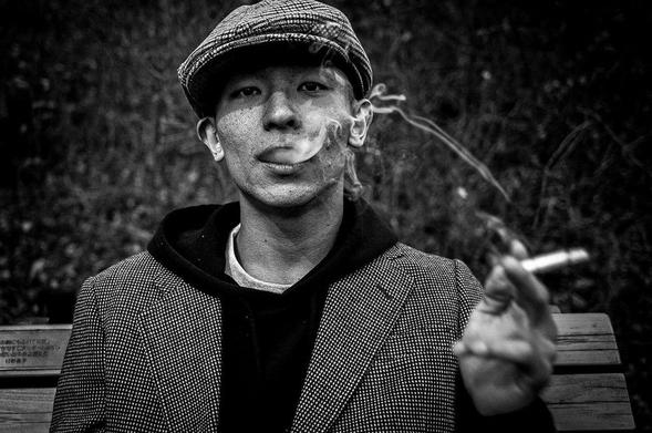 A person in a checkered jacket holds a cigarette in a park, with smoke curling upward, set against a dark, blurred background.