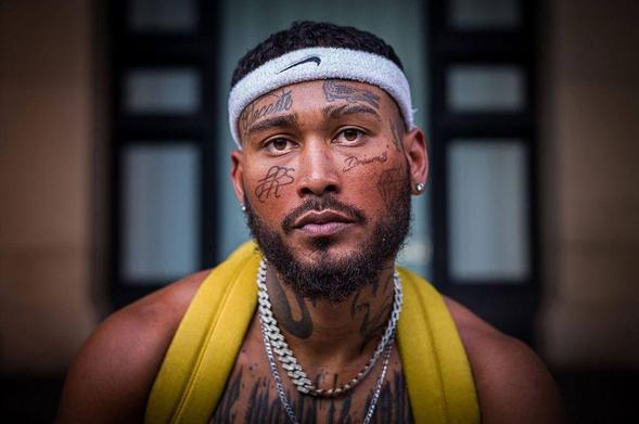 A muscular individual with a yellow backpack, adorned with chains, poses with a confident expression against a blurred background.