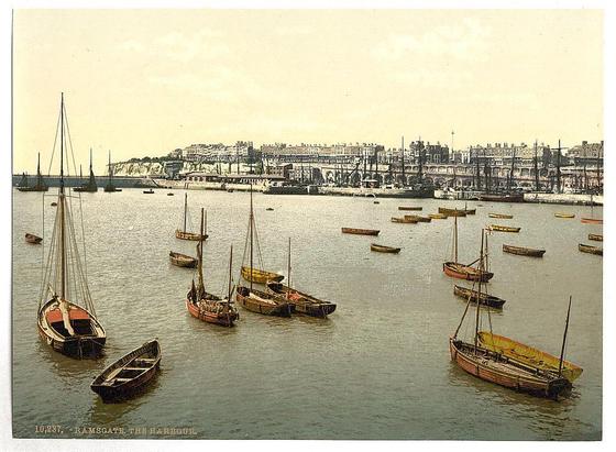 The image depicts a bustling harbor scene, possibly from late 19th to early 20th-century England. The foreground is dominated by various small boats of different sizes and colors gently bobbing on the water's surface. Some have masts raised while others are flat-bottomed with oars or rowing implements visible inside their hulls.

The middle ground reveals a larger vessel, identifiable as a sailing ship due to its tall mast standing prominently amidst smaller wooden boats. The ships seem well-maintained and painted in hues of red, yellow, green, indicating both functional use and aesthetic considerations for the owners or captains who may be local fishermen or merchants traveling between ports.

In the background, an extensive harbor infrastructure unfolds with a series of cranes and docks extending into the water on one side. This industrial backdrop includes several large ships docked at these facilities, likely engaged in loading or unloading goods—a common sight in commercial harbors during that era.

Beyond this human-made landscape rises a cliff adorned with houses and buildings indicative of Ramsgate town's coastal setting. The architecture is characteristic of the period—multistory residential structures with flat facades, some featuring ornate details typical for British seaside towns' housing styles from that historical epoch.

The sky above is overcast, suggesti [...]