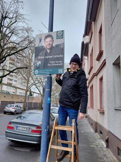 Eine Person in winterlicher Kleidung steht auf einer Leiter und befestigt ein Wahlplakat für Brian Junker-Latocha an einem Laternenpfahl in einer Wohnstraße. Das Plakat zeigt ein Foto von Brian Junker-Latocha mit dem Slogan "Viele Storys. Ein Offenbach!" und einem QR-Code. Im Hintergrund sind ein geparktes Auto, ein Mehrfamilienhaus und kahle Bäume zu sehen.