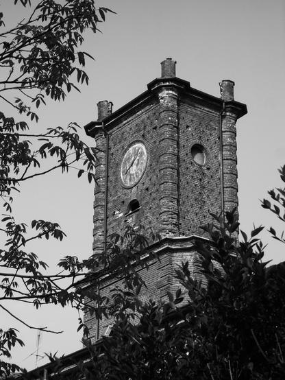 Foto in bianco e nero di una torre campanaria in mattoni rossi con un orologio ormai non più funzionante. La torre non ha la copertura a punta.