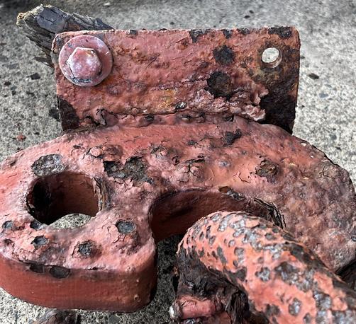 A red rusted metal plate with a bolt and fragment of wood still attached lies on gray concrete. The plate has a red with black pitted anchor chain hook still holding one corroded and pocket marked link of anchor chain. Photo by Robert Emond.