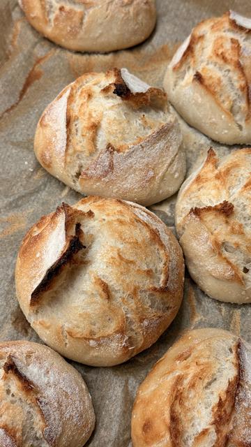 Das Bild zeigt ein Backblech mit mehreren frisch gebackenen Brötchen.  
Die Brötchen sind rund, etwas unregelmäßig geformt und haben eine stark aufgeplatzte Kruste. Die Oberfläche ist goldbraun gebacken, mit helleren, fast weißen bemehlten Stellen und dunkleren, leicht gebräunten Rissen und Spitzen, wo der Teig beim Backen aufgerissen ist. Man sieht die typische, knusprige Struktur einer dünnen Kruste, darunter wirkt das Innere weich und luftig. Das Blech ist mit Backpapier ausgelegt, auf dem sich einige braune Mehl- und Backspuren befinden. Insgesamt wirkt es wie ein Blech frisch aus dem Ofen mit knusprigen, handgemachten Brötchen.