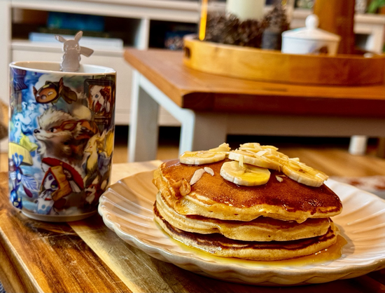 A pile of pancakes with banana slices and golden syrup. A winter-themed pokemon cup of coffee next to it.