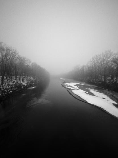A foggy black and white image of a river with snow along its banks and leafless trees on both sides, creating a tranquil and mysterious atmosphere.