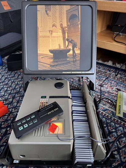 Abgebildet ist ein Diaprojektor, mit dem ein Foto auf eine Mattscheibe projiziert wird. Ringsrum unwichtige Einrichtungsgegenstände.