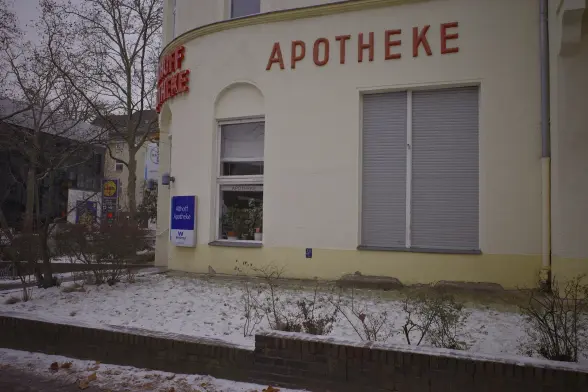 The side of a house with a pharmacy on the ground floor, the walls are yellow with the word "APOTHEKE" (pharmacy in German) in old, red lettering. The shutters on one window are completely down, on the other just halfway. In the foreground a bit of greenery around the house covered in a dusting of white snow.