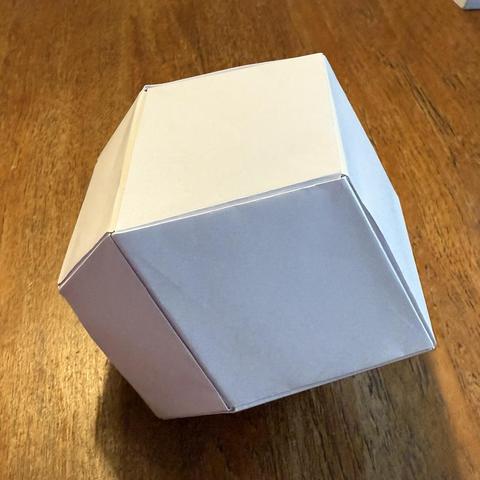 A rhombic dodecahedron made from folded paper. 6 sheets of white A4 paper were trimmed into 12 sheets of A5, after which each sheet was folded into a rhombus with 1:√2 height:width proportions, with alternating sticking-out tabs and pockets into which tabs could be inserted. These 12 rhombi were then assembled into a rhombic dodecahedron, a regular 12-sided shape where each side is a rhombus.