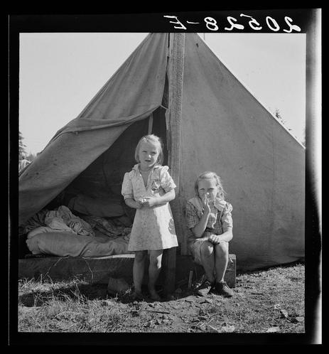 The image depicts a black and white photograph of two young girls standing in front of a tent. The older girl is sitting on what appears to be a crate, while the younger one stands with her hands clasped together. They both wear simple dresses typical of mid-20th-century children's fashion. Their expressions suggest they are accustomed to hardship; their surroundings—a makeshift campsite—reinforce this impression.

In front of them is an open tent revealing some bedding and clothes, indicating a temporary dwelling situation. The ground appears dry with sparse grass, hinting at arid conditions or late summer season in the region depicted. A handwritten inscription "E-8520" overlays the image at the top right corner; this could be a cataloguing code.

The photograph likely documents life for migrant workers during that era, as suggested by additional context provided: it seems to pertain specifically to bean pickers' camps found in agricultural regions of Oregon. The general caption number mentioned implies there are more photographs documenting similar living conditions and circumstances.