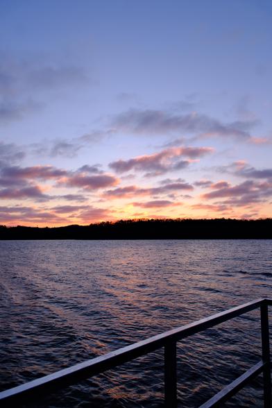 The blue sky and clouds turn orange as the sun rises over the woods on the other shore of the lake. At the bottom is a metal rail of a jetty, and lake water in the middle.