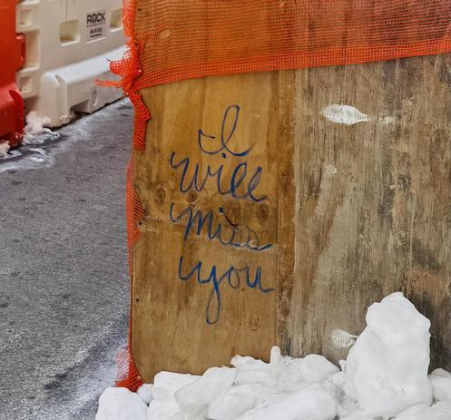 A color photo of the side of a construction site covered in old wooden boards. The right side of the photo is a plain wooden board and the left side of the photo show shows the street and some white and orange bollards. The middle of the photo is a wooden board and someone has written in script with a black marker enlarge letter letters "I will miss you" It made me a little sad.