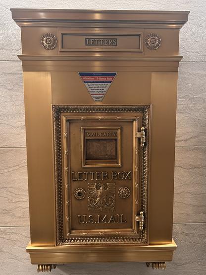 A vintage gold-colored letterbox with the words "LETTERS," "LETTER BOX," and "U.S. MAIL" prominently featured. It includes a decorative design and a warning sign regarding a 13-ounce rule for mail.