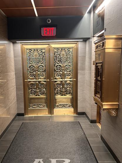 Image of an elegantly decorated elevator entrance featuring ornate gold doors with intricate designs. An illuminated "EXIT" sign is above the doors, and there is a decorative mailbox on the right. Dark tiled flooring complements the luxurious aesthetic.