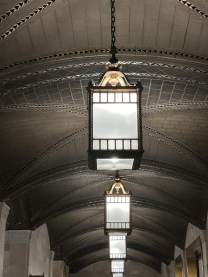A series of ornate hanging lanterns illuminate a grand, arched ceiling featuring detailed architectural patterns and textures. The lanterns are designed with a vintage aesthetic, showcasing intricate details and glass panels.