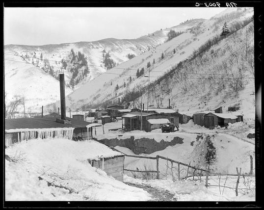 The image depicts a bleak and snowy rural landscape, likely taken during winter. The ground is covered in snow with icicles hanging from the eaves of structures. A collection of dilapidated wooden cabins or shacks are scattered across the scene on sloping terrain leading up to tree-covered hillsides. Some buildings have visible chimneys emitting smoke, suggesting they may still be occupied despite their poor state.

The setting appears impoverished and desolate, with a stark contrast between the natural landscape and human-made structures. The absence of modern amenities or signs of economic prosperity is notable in this otherwise serene winter scene. It conveys an atmosphere of hardship associated with rural living during that period.