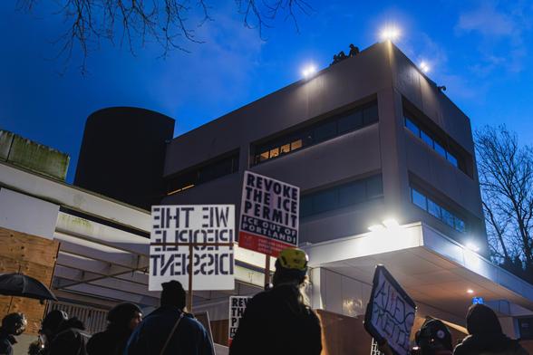A photo of protesters holding signs including "REVOKE THE ICE PERMIT - ICE OUT OF PORTLAND!" and gathered in front of a large building around dusk, with three figures silhouetted against the sky visible on the roof of the building looking down at the crowd.