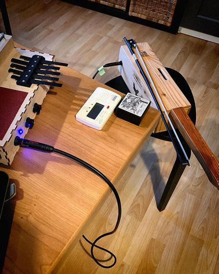A Leaf Audio Sound box (small wooden box, featuring various soundmaking elements: metal springs, a "Kalimba" and a rough surface for scratching) sits on a table, and a DIY one-string violin on a smaller stool next to it. Also on the table: a tuner and violin rosin.