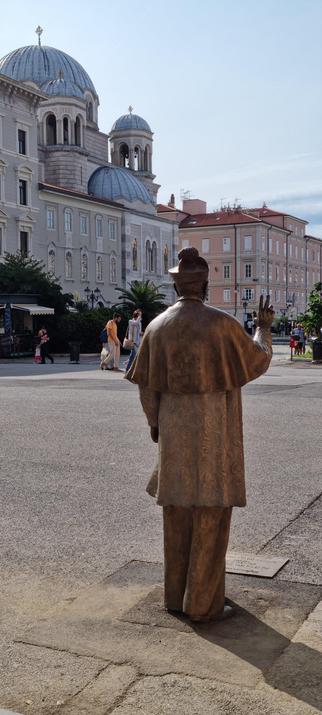 Die Statue von Antonio Santin, von hinten gesehen. Sie zeigt einen Mann im Priestergewand und Hut, der mit seinen Fingern ein Zeichen macht. Im Hintergrund ist die serbisch orthodoxe Kirche zu sehen.