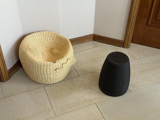 A basket on the floor with a face and little knobs as arms has a giant cat shaped dent in its head while it looks like its reaching out to an upturned wastepaper basket.