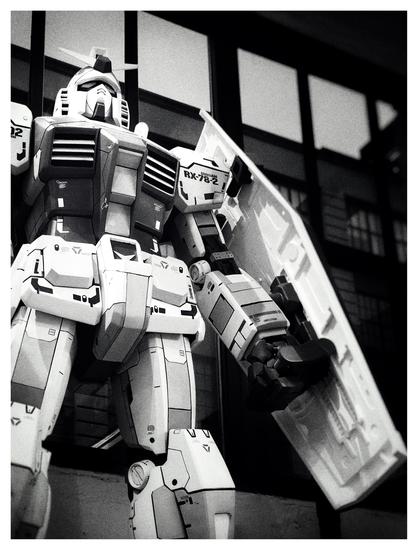 Black and white photo of a large model of a robot from the Gundam anime franchise, seen from the legs up, shot from below looking slightly upwards. The robot stands erect in a protective pose, its left (viewer’s right) fist clenched and carrying a long hexagonal shield. Its other arm is cropped from view. In the background are shadowy window panels with a view of buildings and the evening sky outside.