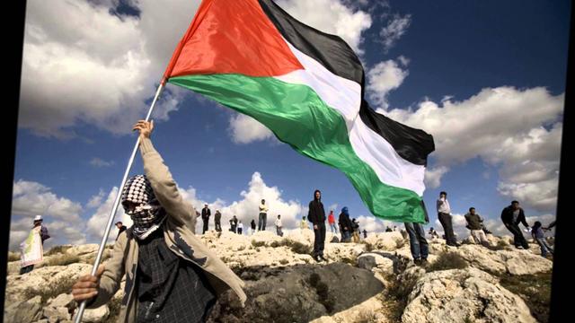 palestine people on a rocky terrain. The one in the foreground is waving a Palestinian flag.