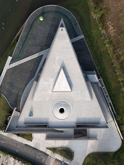 Aerial view of a modern, angular concrete building with a central circular opening and a green accent near a waterway.