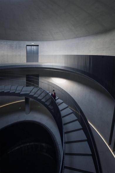 A spiral staircase with smooth concrete and metal railings, featuring a person walking down the stairs, illuminated by soft lighting.