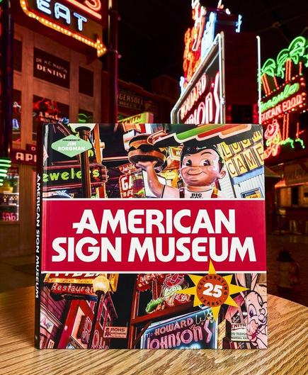 Cover of a book that says "American Sign Museum" in a horizontal band across the centre. Above and below this are scenes from the museum with a variety of brightly coloured and illuminated signs. The book is stood up on a wooden table with some of those sign scenes in the background.