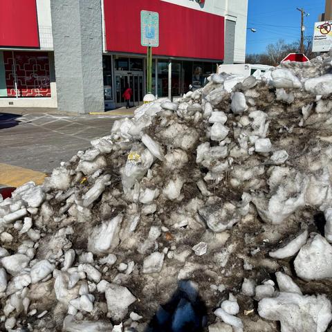 A large pile of dirty, chunky ice and snow completely fills a handicapped parking space in front of a CVS pharmacy. The ice pile is several feet high and extends across the entire designated parking spot, making it completely inaccessible. A blue handicapped parking sign is visible on a post nearby. The CVS storefront has red and gray exterior walls with large windows, and there's a person in red clothing walking near the entrance.