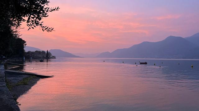 Ein ruhiger See bei Sonnenuntergang. Die Wasseroberfläche spiegelt den rosafarbenen und lilafarbenen Himmel wider. Im Hintergrund erheben sich sanfte Berge, die in der Abenddämmerung silhouettiert sind. Am Seeufer sind einige Gebäude und Bäume zu erkennen. Ein kleines Boot ist auf dem Wasser zu sehen. Die Stimmung ist friedlich und idyllisch und verbreitet Ruhe und Zufriedenheit.
