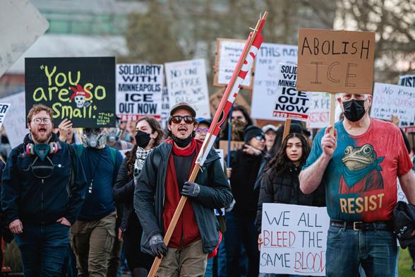 A photo of a dense crowd of protesters marching outside around sunset wearing face masks and carrying various signs including "YOU ARE SO SMALL" with an illustration of a skull wearing a red jester's cap that says "MAGA" on it, "ABOLISH ICE," and "WE ALL BLEED THE SAME BLOOD." One man wears a red and blue T-shirt with an image of a Portland resist frog on it and the word "RESIST" underneath it.