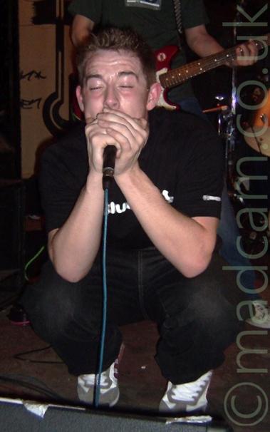 The singer from a band, crouching down on stage and singing into a microphone held in his hands, his eyes closed.
He is wearing a black t-shirt and black jeans, and has short, spiky brown hair
A man wearing a green t-shirt and playing a red and white guitar can be seen in the background