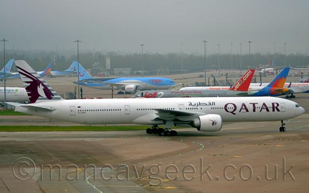 Side view of a twin engined jet airliner taxiing from left to right at a busy airport.
The plane is largely white, with mauve, billboard-style "Qatar" titles on the forward fuselage, repeated in a grey Arabic script on the upper forward fuselage, above the wing root, and the black registration "A7-BOF" on the lower rear fuselage, just below the last few cabin windows, and a small Qatari flag just above those windows.
The tail is a very light grey, with darker grey stripes overlaid with the head of an animal with horns sticking out of the back of it's head.
A smaller, circular version of the tail logo appears on the side of the white engine pods mounted under the wings.
Grey concrete apron, marked with yellow lines for planes to follow, some of which are lit up, fills the foreground.
More apron space fills the background, with a total of 13 jet airliners visible, parked facing in various directions, in an array of liveries that predominantly feature shades of white, blue, grey, and red.
Trees in the distance are  being swallowed up by grey mist.