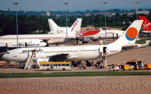 Side view of twin engined jet airliner parked facing to the left.
The plane is almost entirely white, with black "PremiAir" titles on the upper forward fuselage, including orange and blue dots over the lower-case letters "i", and the registration "OY-CNL" on the lower rear fuselage in front of the rear passenger door, as well as black text "Airbus A300" after the door.
There are also large orange and blue circles on the tail, the orange one partially overlaid on the blue one.
The "PremiAir" logo is repeated in a smaller form on the sides of the engine pods under the wings.
A white bus with a yellow roof is parked by the engine pods, and another under the planes rear fuselage, with a snake of people walking towards the latter, mostly coming from the airstairs connected to the open rear cabin doors, but also some walking past the other bus.
A strip of green grass separates the plane from a large area of concrete apron that fills the foreground, with more apron in the background filles with 4 more large jet airliners parked facing in various directions, all in various shades of white, blue, and red.
Tall lighting poles amongst them reach into the sky, with trees and buildings stretch off into the distance, meeting grey sky.