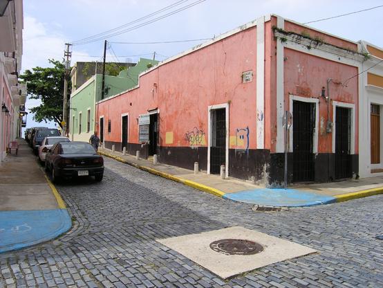There is a rundown pink house on the corner of a small cobblestone intersection. The bottom of the building is painted black, the walls are a dirty pink, and there is white trim around the doors, windows and frame. Graffiti is painted on the walls. Behind the pink building is a pale green building. The streets are very narrow; cars parked on the side of street barely leave any room to pass. Power lines pass over the street and buildings. The sky is overcast, but shadows can still be seen. San Juan, Puerto Rico.