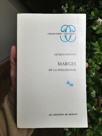 A hand holds a copy of the book "Marges de la philosophie" by Jacques Derrida.