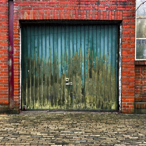 A textured green metal door with streaks of rust and moss is set against a backdrop of red brick walls. The door features vertical panels and a simple lock. The ground is made of cobblestones, adding to the rustic atmosphere.