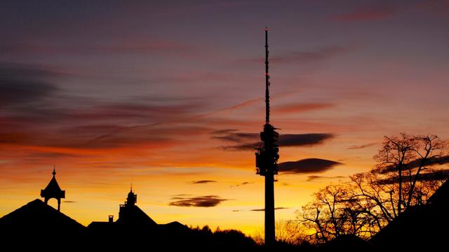 Das Bild zeigt eine Silhouette von Gebäuden und Bäumen vor einem leuchtenden Sonnenaufgang. Im Hintergrund befindet sich ein hoher Sendemast. Der Himmel ist in warmen Farben wie Orange, Gelb und Rot gehalten, die Wolken sind in verschiedenen Schattierungen dieser Farben. Die Gebäude und Bäume sind dunkel und heben sich deutlich vom hellen Himmel ab. Das Bild vermittelt ein Gefühl von Ruhe und Schönheit der Natur.
