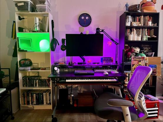 Same white shelf as before on left side. Dark brown desk in middle. Midi keyboard underneath. 27" Monitor on hydraulic VESA arm. Headphones hanging from left side of monitor. Two MacBook airs sideways. Silksong Hornet figure. Super Metroid themed switch stand. Black bookcase to right. Clock guy from 'Don't Hug Me I'm Scared' on wall above monitor.