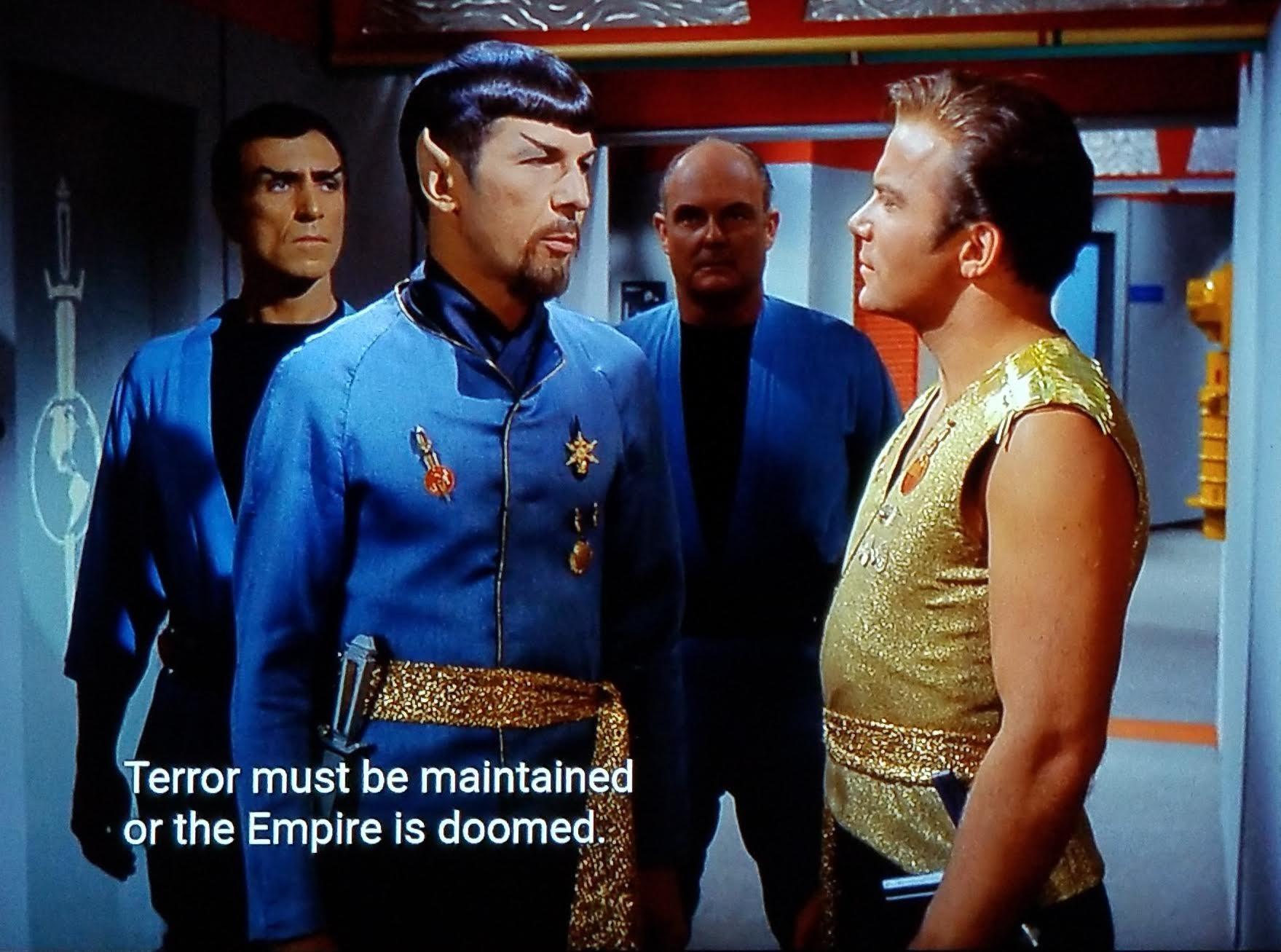 TOS scene, we're in a corridor on the Enterprise. Two random crew members are standing behind Spock in a bodyguard stance. (Tough guy bodyguard vibe, not Coster/Whitney bodyguard vibe). Spock is wearing a dress uniform with extra medals and pins, a golden sash, and has a goatee. He's looking at Kirk, who seems a bit cocky and is wearing a glittery gold, sleeveless uniform. Closed caption reads, "Terror must be maintained or the empire is doomed."