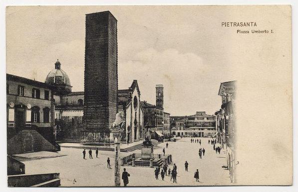The vintage postcard depicts a historical scene from Pietrasanta, specifically Piazza Umberto I. The focal point is the Torre Pisana, an imposing tower rising above surrounding buildings and structures in the square. This architectural feature stands as one of Italy's notable landmarks.

Adjacent to the tower is what appears to be Santa Maria della Quercia church with a prominent dome visible on top. Other significant edifices frame this public space: classical facades, arched entrances, and windows that suggest a bygone era in terms of architecture. The square itself has an open-air feel; people are seen walking or standing around, giving life to the historical ambiance.

The image captures black-and-white photography typical for its time period, likely early 20th century based on clothing styles observed, such as long coats and hats worn by individuals, both men and women. The postcard bears a caption "PIETRASANTA Piazza Umberto I" in italicized text at the top right corner.

The overall condition of the card shows some signs of aging with creases indicative of handling over time or perhaps folding for mailing purposes. There's no direct indication if any specific events, such as military parades, are occurring; however, it portrays a moment of everyday life within this historic European town square.