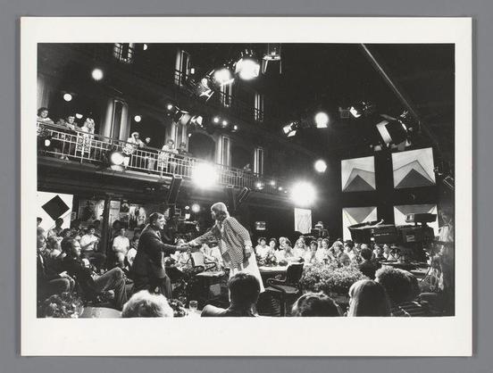 The image depicts a lively scene from what appears to be a television talk show set in an indoor venue, possibly during the 1970s or early '80s judging by the style of clothing and black-and-white photography. A central figure stands on stage addressing a seated audience while being filmed with professional lighting equipment overhead. The individual is dressed casually yet flamboyantly, wearing a striped top and dark pants. Surrounding this person are various other individuals: some standing in what seems like an entrance or balcony area above the main set, possibly crew members; others sitting at tables on stage looking attentive towards the central figure.

The audience below comprises people of different ages and genders dressed semi-formally to casually, with several engaged directly with the performer. The environment is bustling yet orderly, indicative of a public event where media coverage is present—evidenced by microphones in front of some seated attendees suggesting they might be guests or participants being interviewed.

In terms of lighting, there's a mix of ambient and artificial light that focuses on both the stage area and individual audience members. The set includes elements like banners with text possibly indicating show names or sponsors but are not entirely legible from this angle. Overall, it captures an energetic atmosphere typical of live televisio [...]