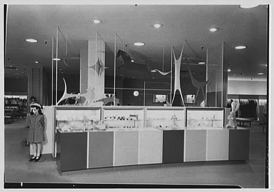 The image depicts a vintage scene inside the toy department of Lord & Taylor, a business located in Westchester. A young child stands near an exhibit featuring various toys and games displayed on shelves within glass cases. The background showcases mid-century modern decor with geometric forms adorning the walls above the display case. Overhead lighting illuminates the space. This photograph is labeled as part of the Gottschalk-Schleisner collection, which consists of one safety film negative measuring 5x7 inches in size.