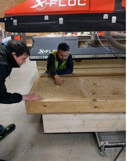 People at bioblow looking at a wood panel filled with chopped straw by a blow in hood on a semi automated wood frame production line